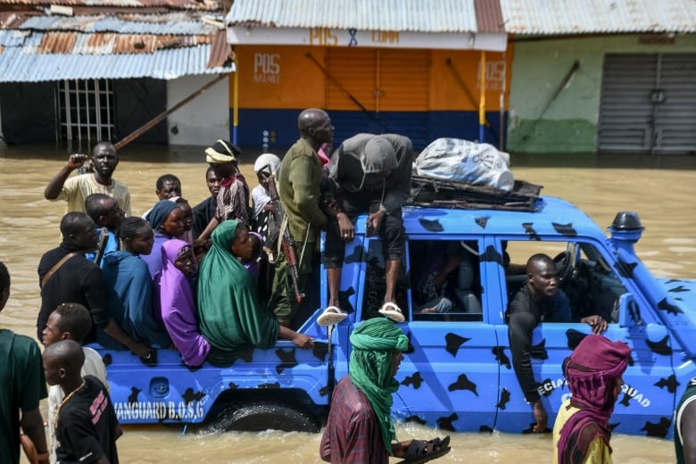 People packed into the bed of a blue truck with its tires almost completely submerged in murky water.