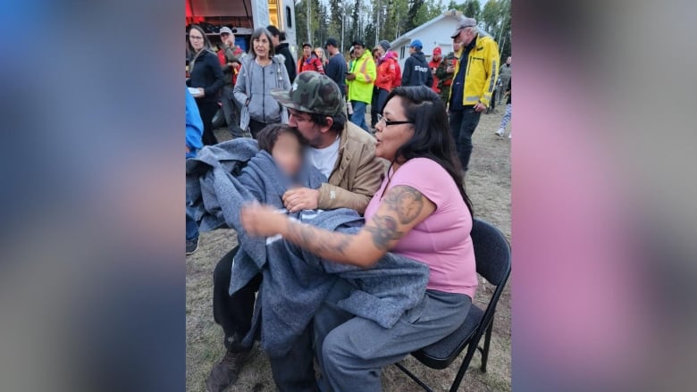 Two parents sit in camp chairs in an outdoor area. The father is kissing his child who is in his arms wrapped in a blanket. The mom holds on to the blanket while a crowd of people look on.