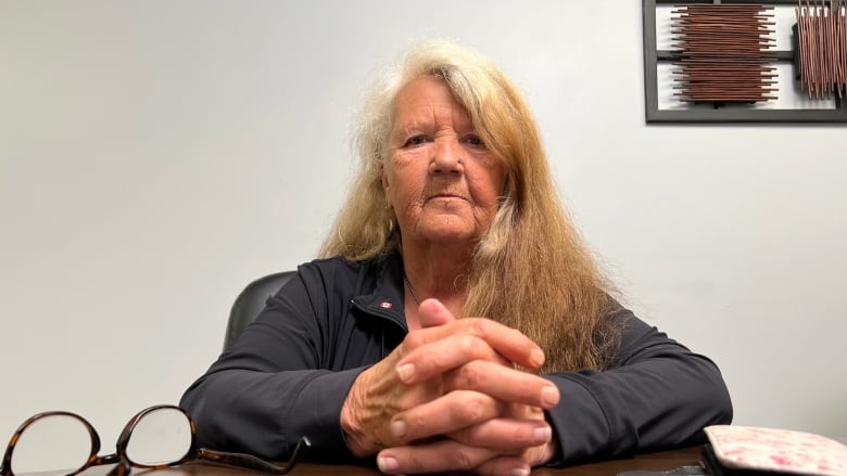 A portrait of a woman with long hair and dark shirt. Beside her is a cell phone and a pair of eyeglasses. 