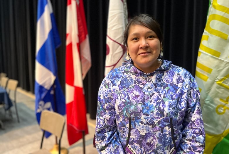 A woman in a purple floral hoodie poses in an official room with the Canadian and Quebec flags behind her. 