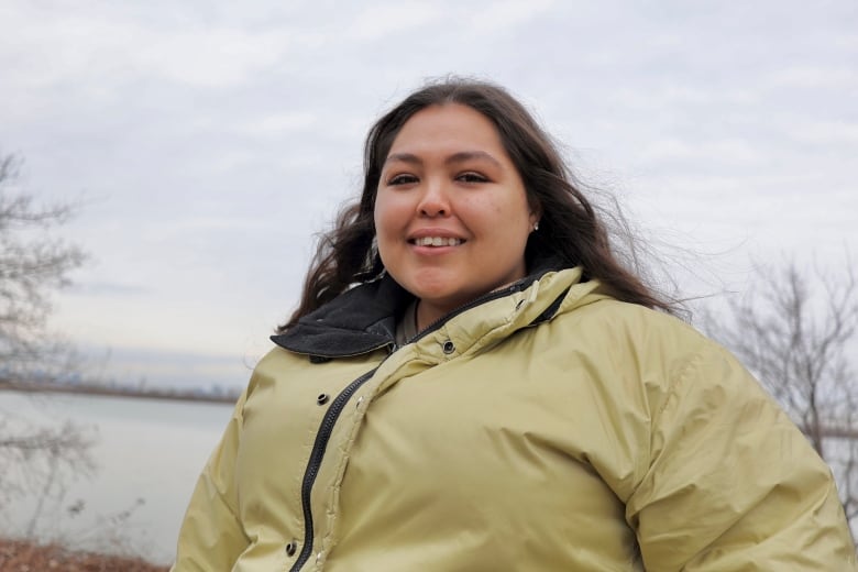 A woman in a green raincoat poses in front of a river. She's smiling and has long brown hair. 