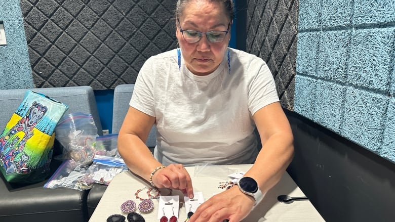 A woman is shown seated at a small table arranging several pairs of multicoloured beaded earrings.