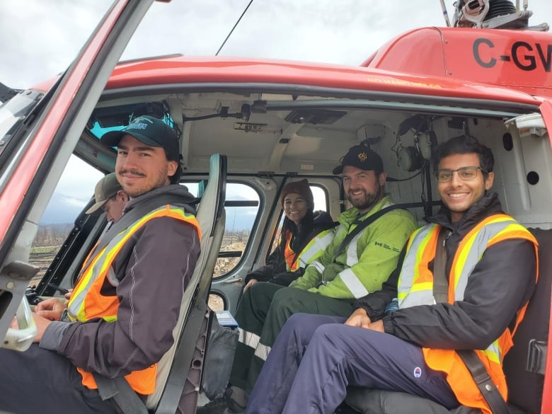 Five people wearing safety vests sit inside a helicopter on the ground.