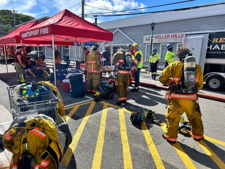 A number of firefighters in their fire gear are on a town street beneath red tents. 