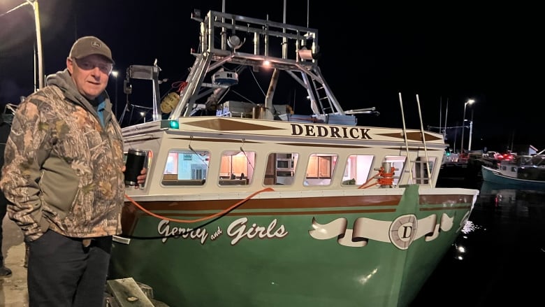 A white man is standing on a deck beside a boat at harbour. He is wearing a camo sweater and a ball cap. The boat beside him reads 