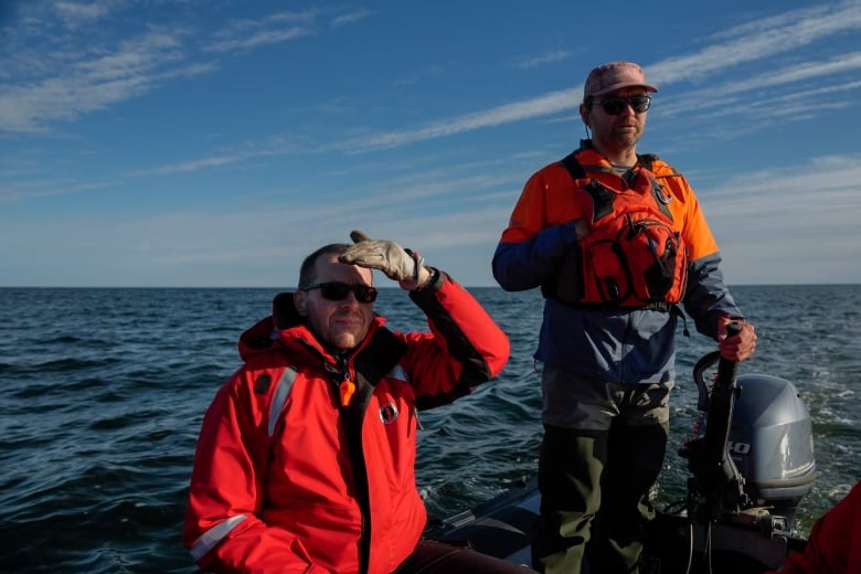 Two men in orange jackets stand on a boat in the open water.