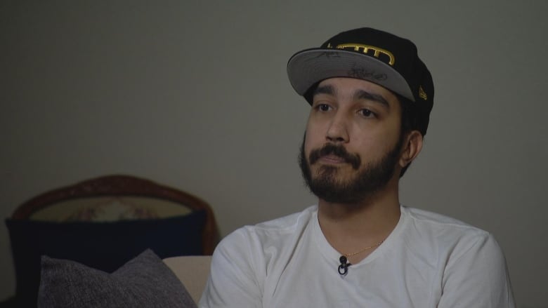 Sallay-Ferreria, a man with a dark beard, wearing a white t-shirt and a baseball cap, sits on a couch in his home.