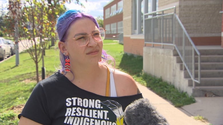 A woman with blue and purple hair speaks into a microphone while standing outside. 