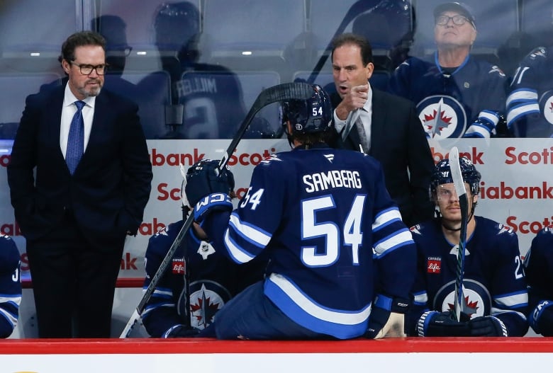 A male ice hockey coach wearing a suit points his right finger while addressing a player on the bench.