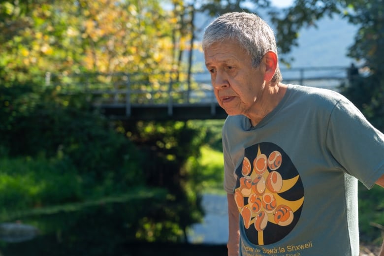 An older Indigenous man wearing a grey T-shirt looks concerned at a waterway below him.