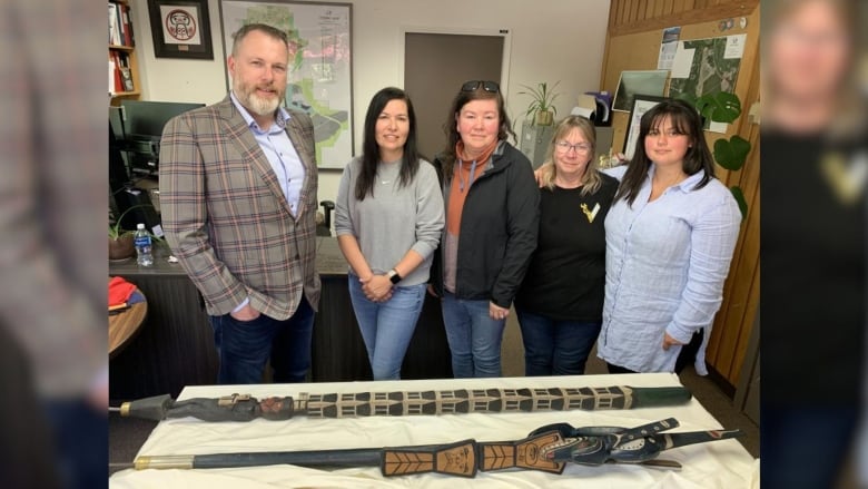 A group of people smile in a municipal council office, with two elaborately designed sticks in front of them on a table.