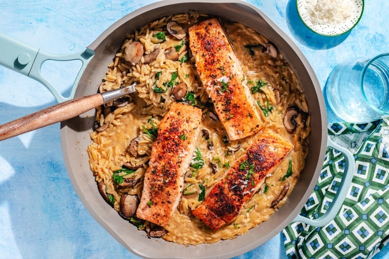 Overhead shot of a pan with creamy orzo and mushrooms with three salmon fillets on top.