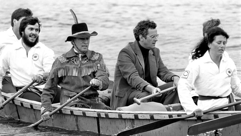 Prime Minister Justin Trudeau is wearing an Indigenous made leather coat and full-brimmed hat as he sits in a canoe and paddles behind former communications minister Francis Fox, who is wearing a black turtleneck and a brown blazer as he holds a paddle in both hands but does not paddle. 