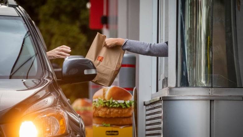 A close-up of a McDonalds employee's arm handing a fast food bag to a customer sitting in their car in a drive-through.