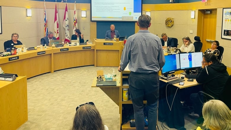 A man, his back to the camera, stands at a podium addressing people sitting at desks with nameplates on their front.