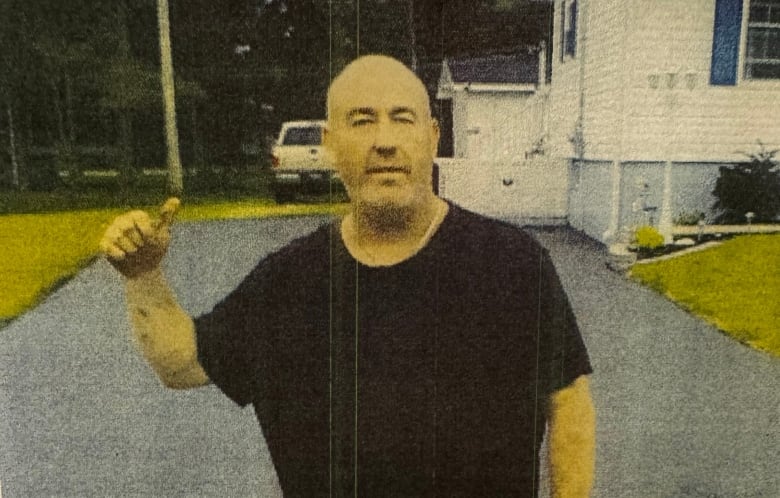 Bald man in black t-shirt standing in a driveway of a home. 