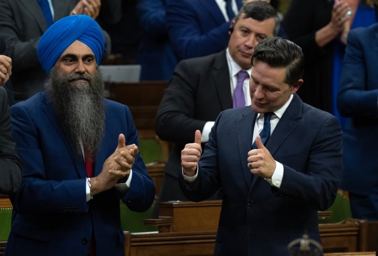 A man in a dark suit gives two thumbs up as he stands in the House of Commons. Those around him applaud.