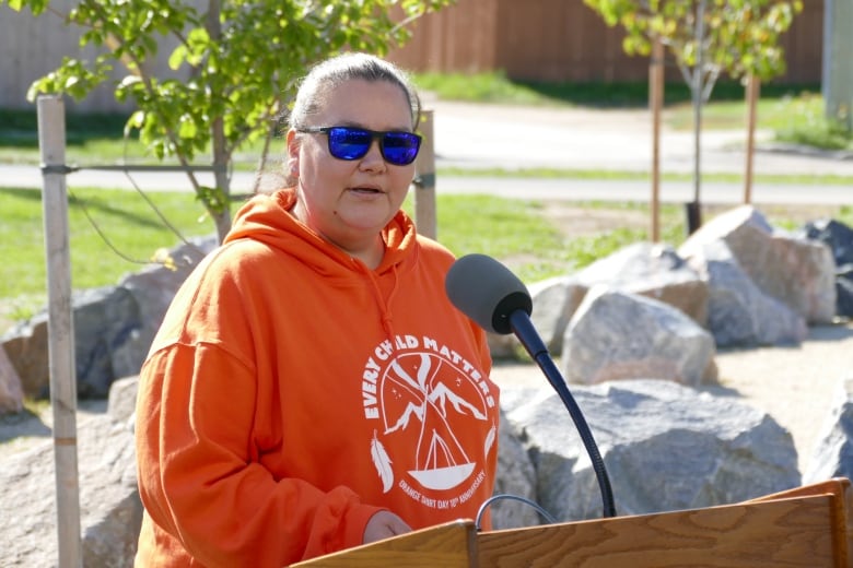 A woman wearing blue sunglasses and an orange hoodie stands at a podium outside. 