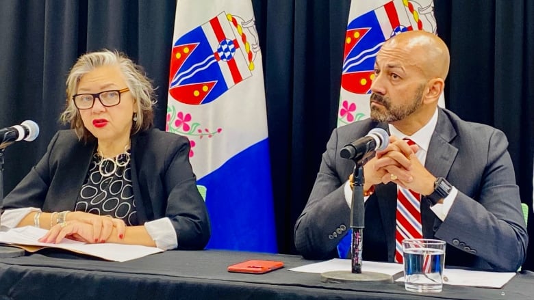 A man and a woman sit at a table behind microphones in front of a dark curtain and some flags.