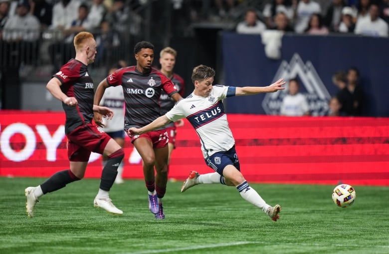 A soccer player bends to kick a ball, as three opponents swarm him from behind.