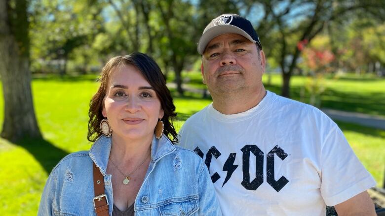 A man , wearing a t-shirt and ball cap poses with a woman, who wears a jean jacket and has her hair down.