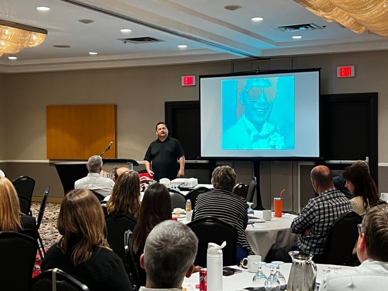 A man wearing a dark collared shirt stands in front of a crown of people to give them a presentation on residential schools.