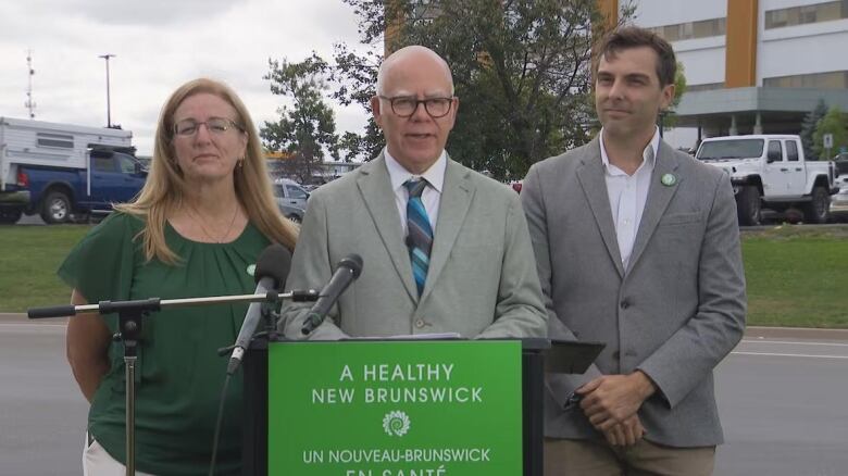 A man in a grey suit stands between a man and a woman outside at a microphone.