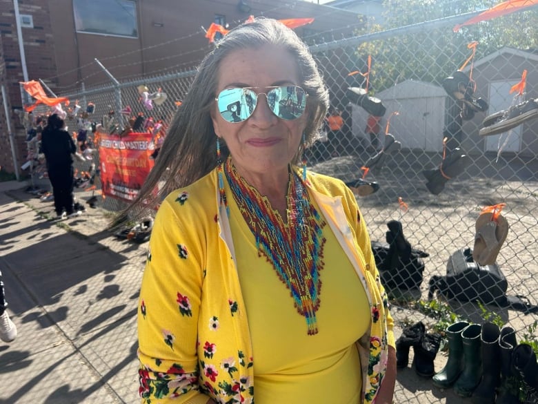 A woman in a yellow outfit and Indigenous jewelery stands in front of a wall of shoes. 