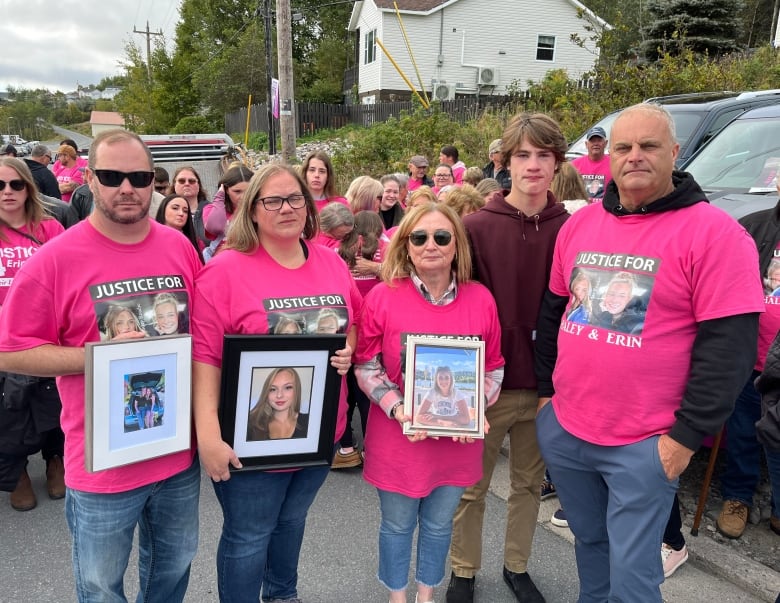 Five people stand next to each other wearing pink shirts holding picture frames of two young girls, crowd of people in pink shirts behind them.