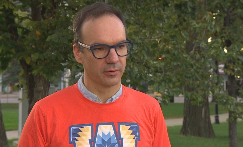 A man with short hair and glasses stands outside near trees. He wears an orange shirt.