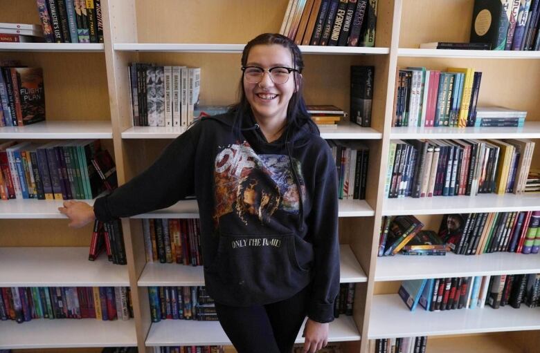 A high school student is pictured standing in front of a book shelf.