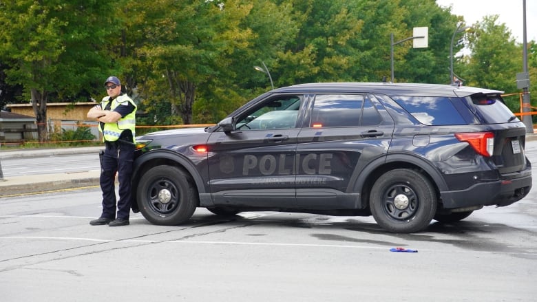 A police car parked behind orange police tape that stretches across a city road on a cloudy day. An officer in a safety vest is standing next to it.