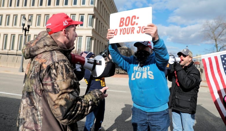 Man in camo and a red MAGA cap stares down a person holding a sign demanding that the election be certified