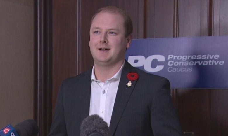 A man with short blonde hair stands in a hallway, in front of a door, and speaks into microphones on a podium.