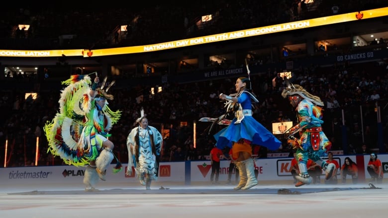 Four people in colourful Indigenous regalia dance on a mat on the ice of a darkened hockey stadium.