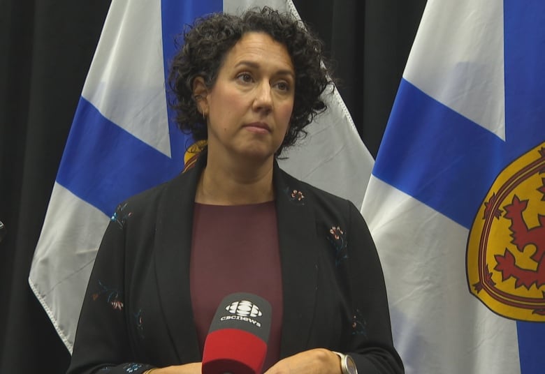 A woman with dark curly hair stands in front of microphones with Nova Scotia flags behind her.