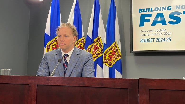 A man sits at a podium with Nova Scotia flags behind him.