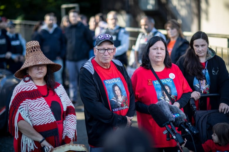 A woman in Indigenous regalia stands beside a man and a woman bearing shirts bearing the picture of a young Indigenous woman with the words, Missing atop the picture and Kristina Walker below it.