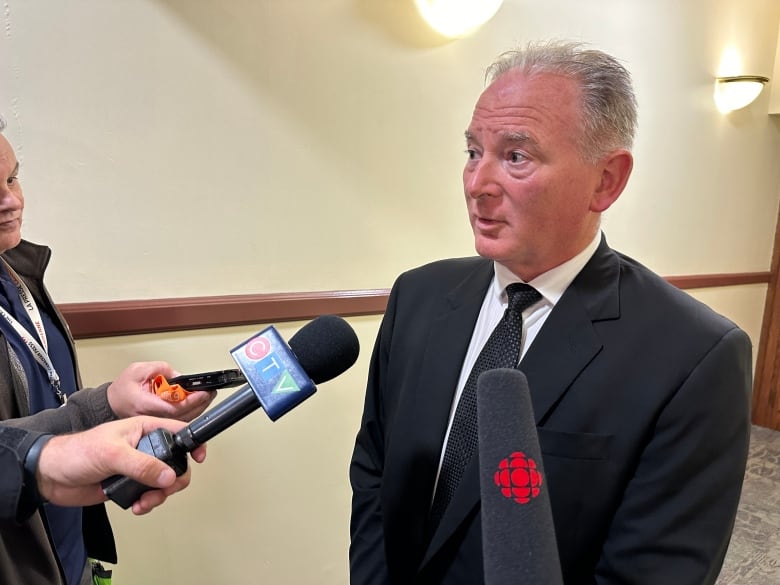 A man in a black suit, white shirt and black tie speaks to reporters who are holding microphones.