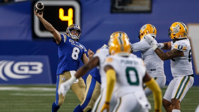 A quarterback in blue throws a football while four opposing players in white look on.