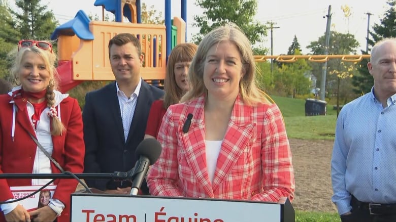 A woman wearing a red blazer, with a group of people and a park behind her.