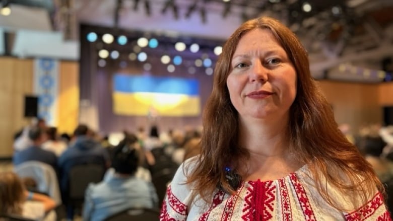 Woman stands and in the background there is a stage and a Ukranian flag. 