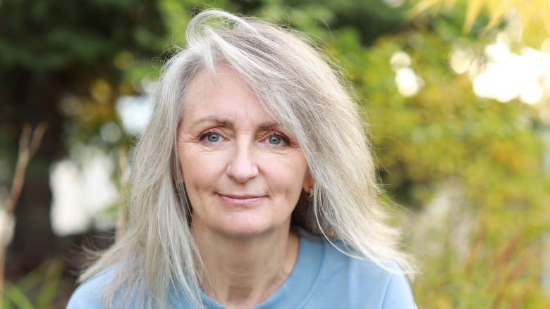 A white woman with long grey hair is crouching outside. She is wearing a light blue screw neck sweater.