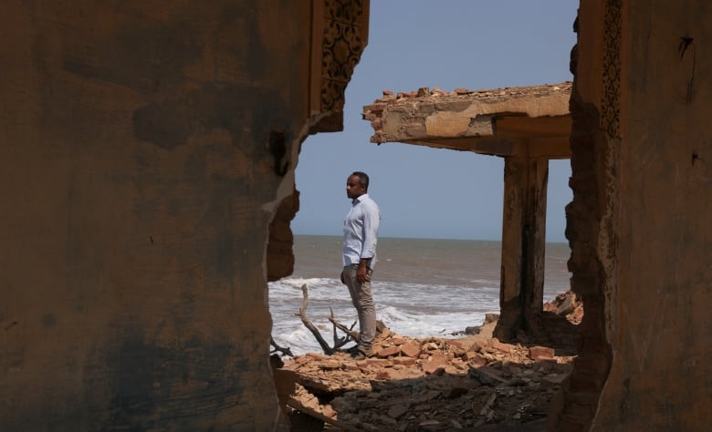 A man stands in the centre of the image, framed by the opening in the rubble of a building. The sea is visible beyond him. 