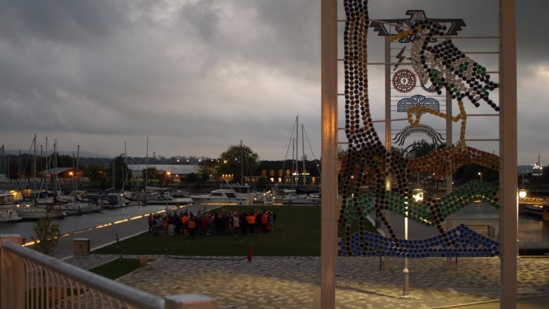 Indigeneous public art is in the foreground, a group of people in orange shirts are in a circle in the middle distance.