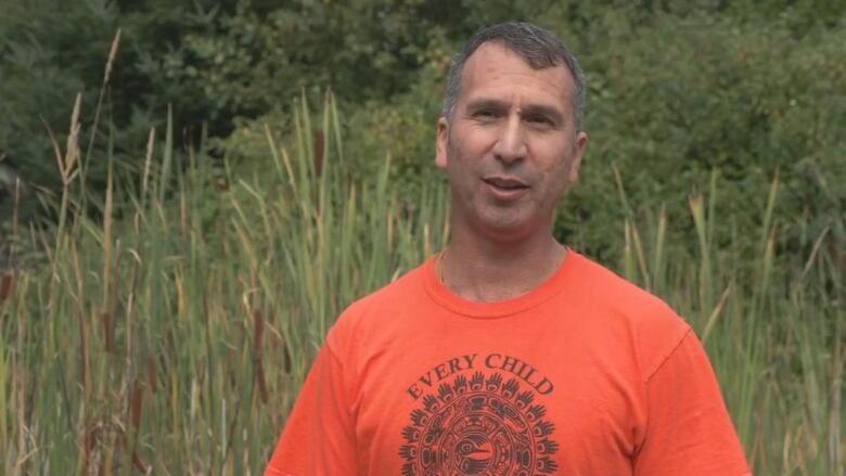 A man wearing orange speaks in front of a green field.