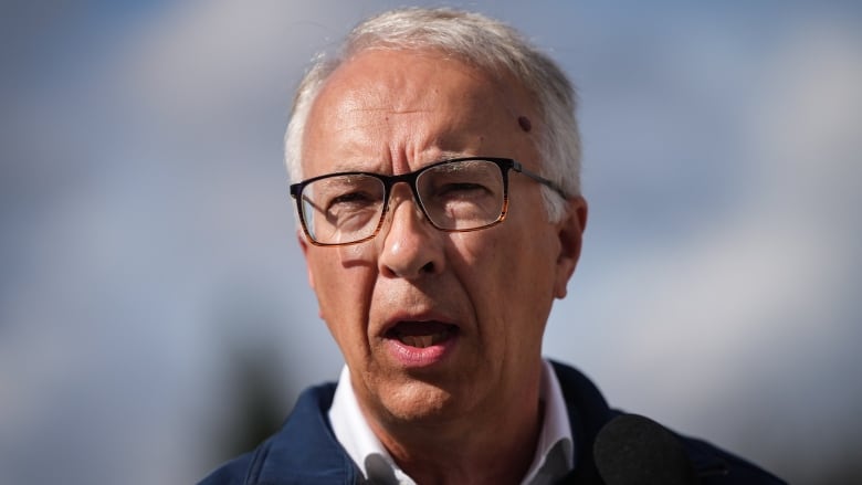 A man with white hair and glasses wearing a blue outdoor jacket over a white open-collared shirt gives a speech.