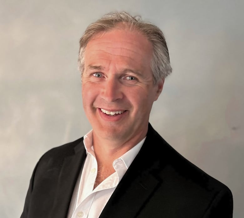 A man in a black blazer, white shirt standing in front of a grey background.