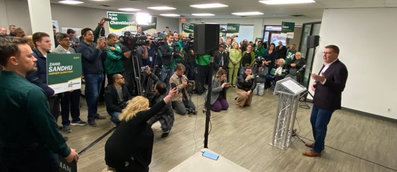 A man in a suit speaks at a podium while a crowd of supporters listens.