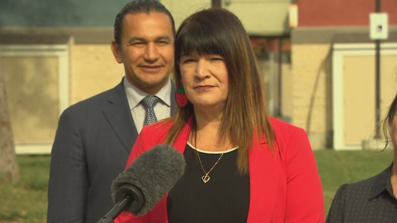 A woman with long brown hair, and wearing a red blazer, stands outside at a podium.